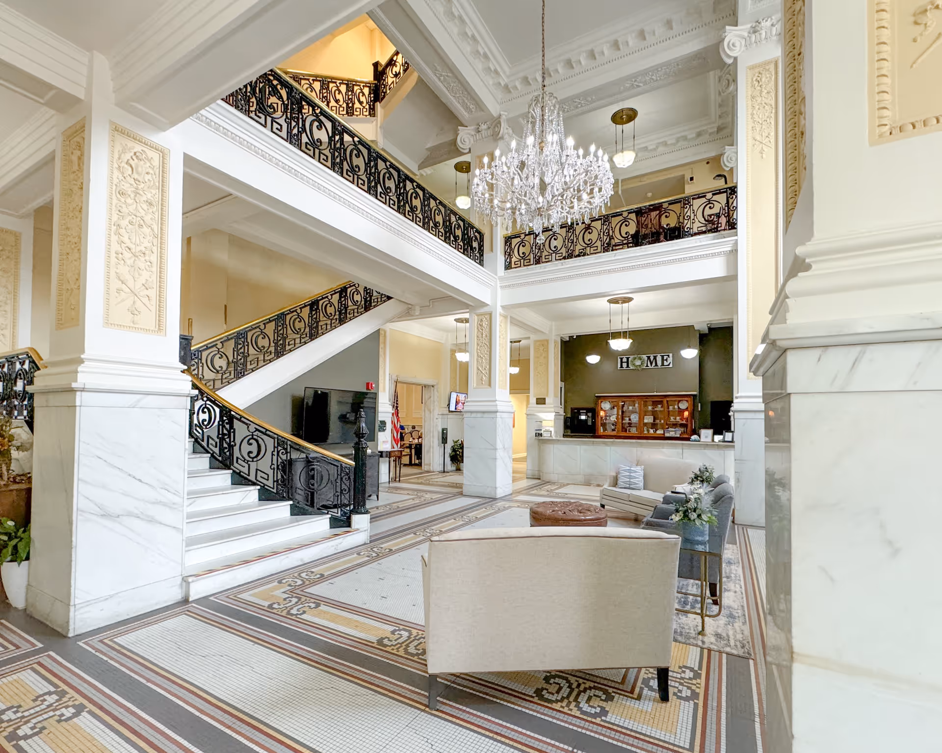Spacious and elegant lobby area with a high ceiling, ornate chandelier, marble columns, and decorative railings on the staircase and balcony. The floor features intricate tile work, and there is a seating area with sofas and chairs. A reception desk is visible in the background with a sign that reads 'HOME'.