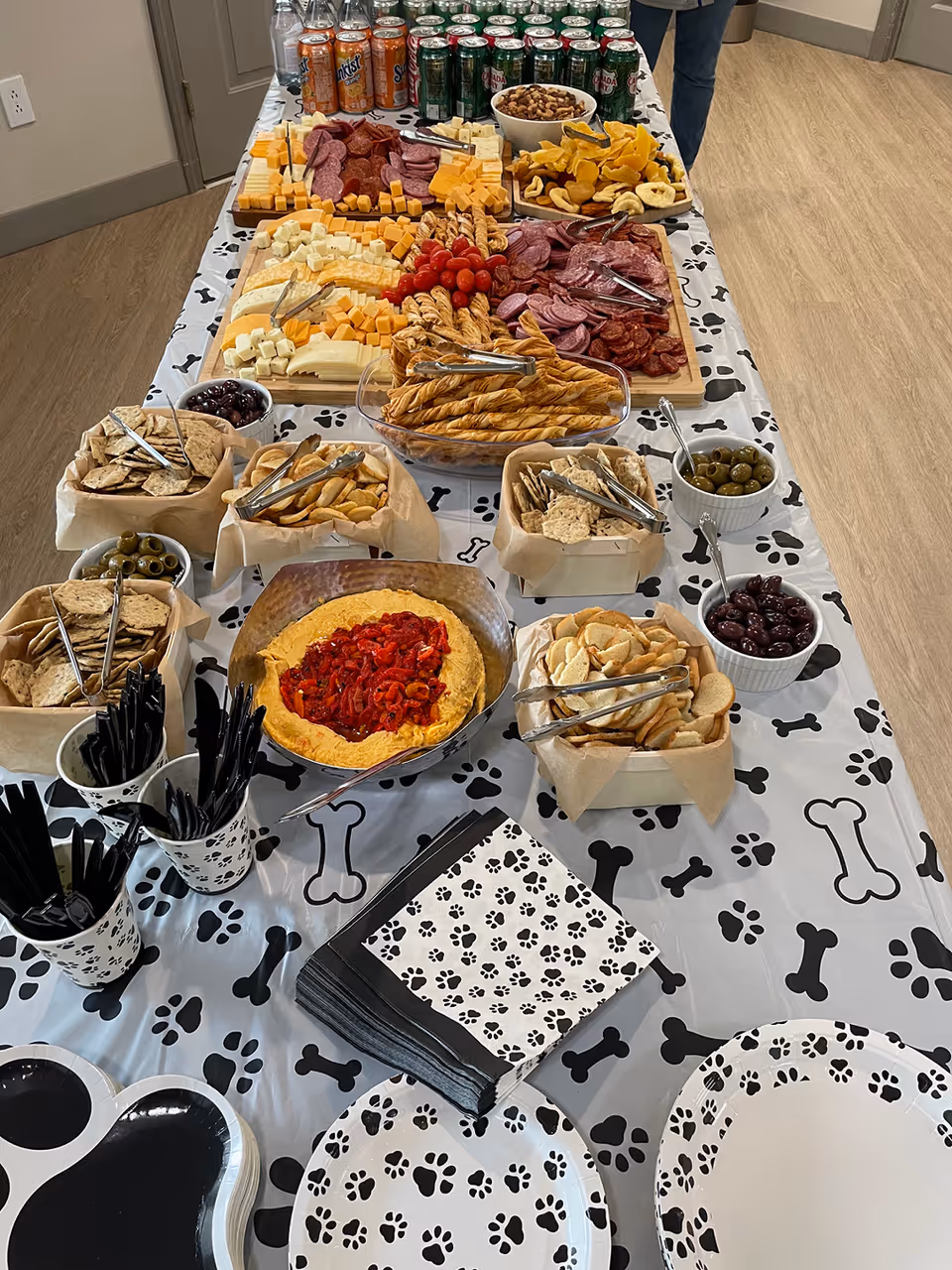 A long table covered with a white tablecloth decorated with black paw prints and bones, set with a variety of snacks including cheese cubes, sliced meats, breadsticks, crackers, olives, and a bowl of dip topped with red peppers. There are also cans of soda and bottled water at the far end of the table, along with black plastic utensils in paw print cups and matching napkins and plates.
