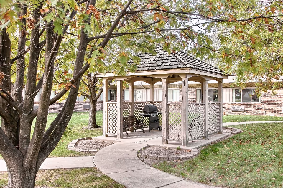 A wooden gazebo with a bench and grill sits in a landscaped courtyard outside a brick building, surrounded by trees and a curved walkway.