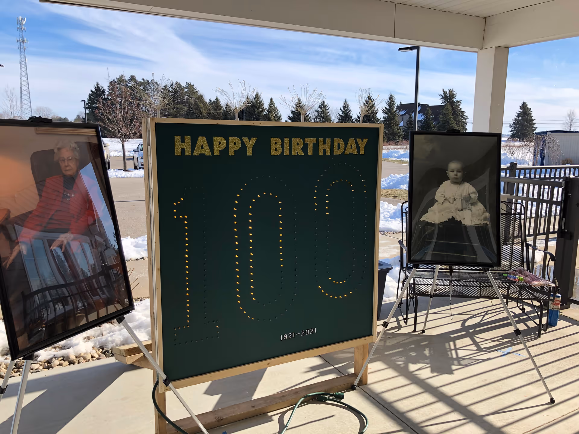 Outdoor covered patio area with a large green sign that says 'HAPPY BIRTHDAY 100 1921-2021' in gold letters and lights. On either side of the sign are framed photos on easels: one of an elderly woman sitting in a chair and the other of a baby in a white dress. Snow is visible on the ground and trees in the background.