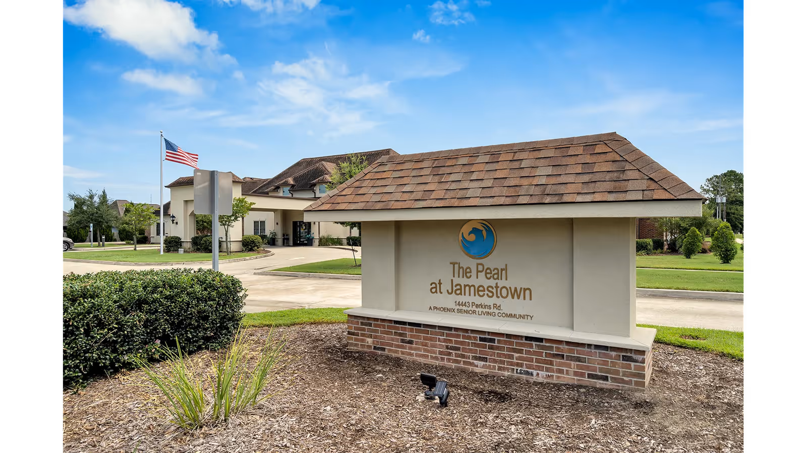Entrance sign for The Pearl at Jamestown memory care community in front of the facility building with an American flag and landscaping.