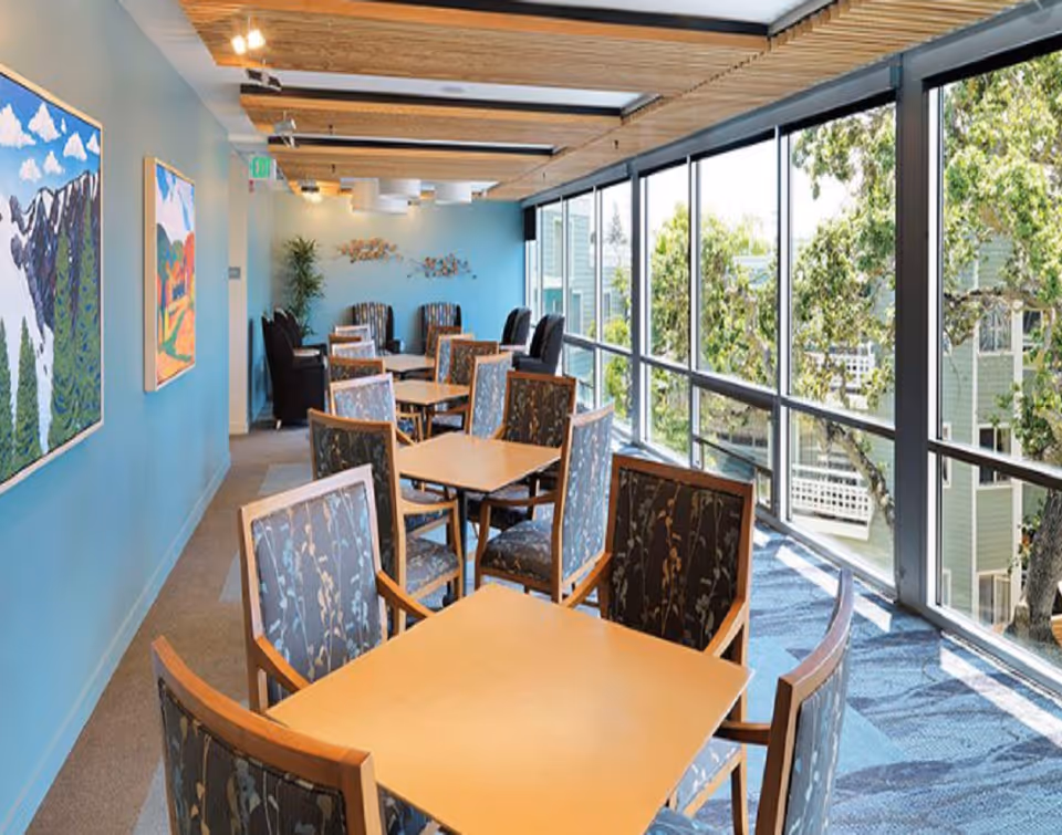 Bright indoor dining area with wooden tables, patterned chairs, and large windows overlooking greenery.