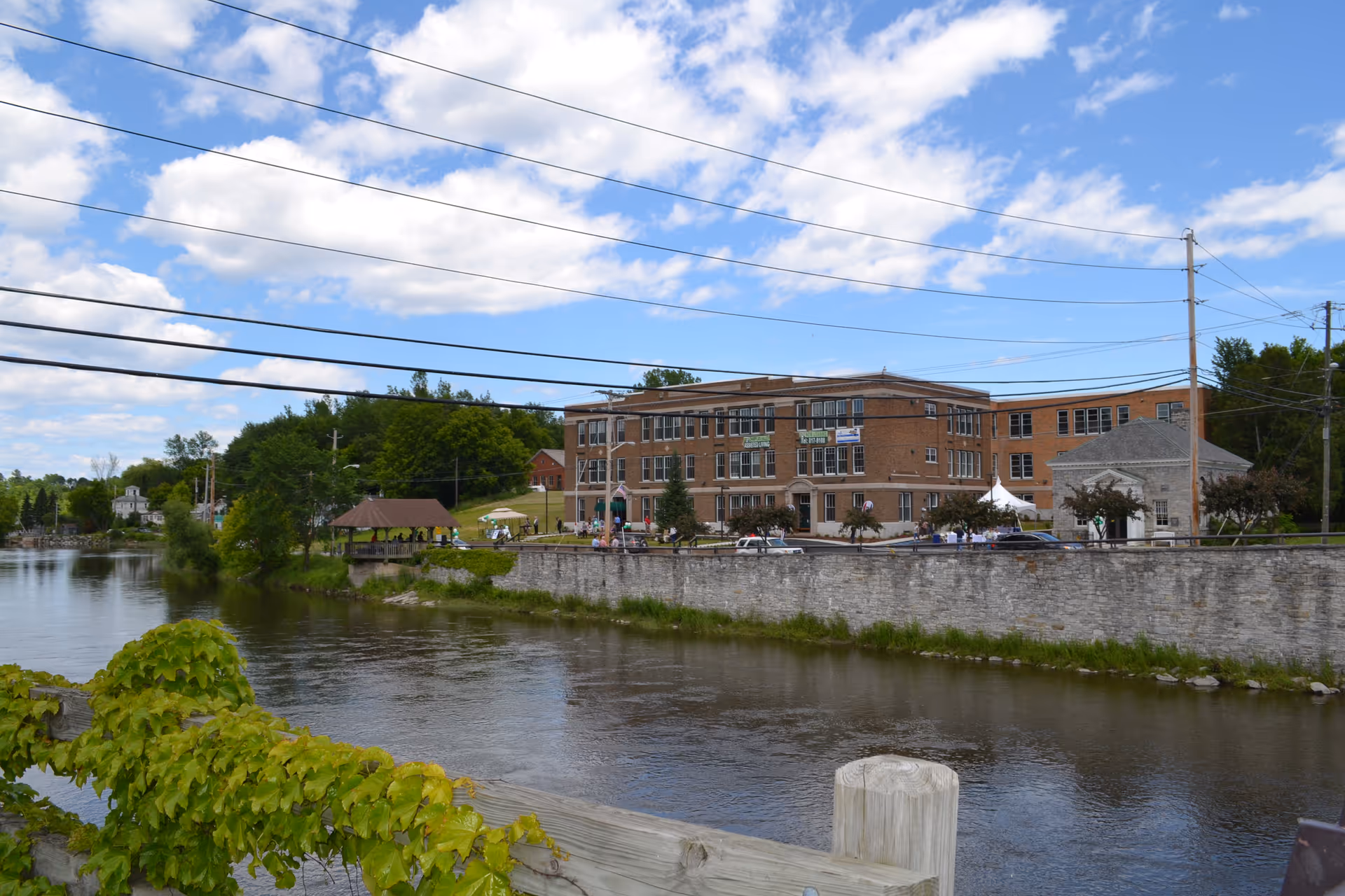 Brick senior living facility building along a river with a stone embankment, gazebo and tents under a partly cloudy sky.