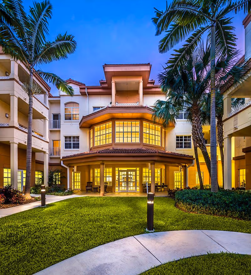 Well-lit three-story senior living building with a landscaped courtyard, palm trees, and illuminated windows at dusk.