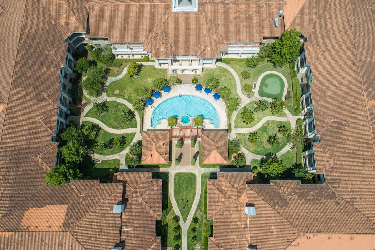 Aerial view of Conservatory At Alden Bridge showing a central courtyard with a swimming pool, surrounding pathways, green lawns, trees, and buildings with terracotta roofs.