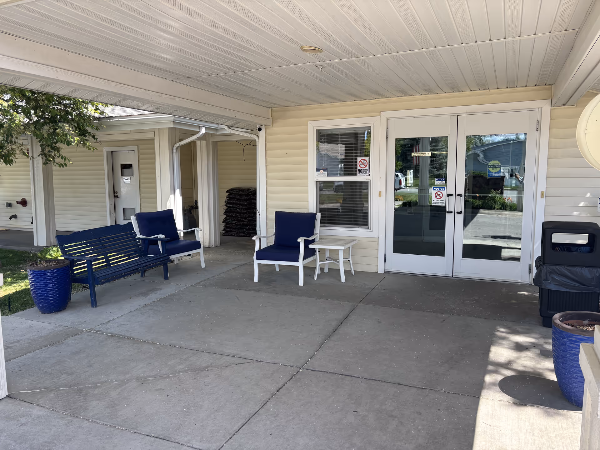 Covered outdoor seating area at Kalispell Assisted Living with two white chairs with blue cushions, a blue bench, two blue planters, a small white table, and double glass doors leading inside the building.