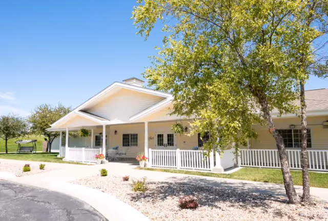 Exterior view of a single-story building with a covered porch, white railing, and hanging plants. There is a tree with green leaves in the foreground and a clear blue sky above. The area around the building has landscaping with rocks and small shrubs.