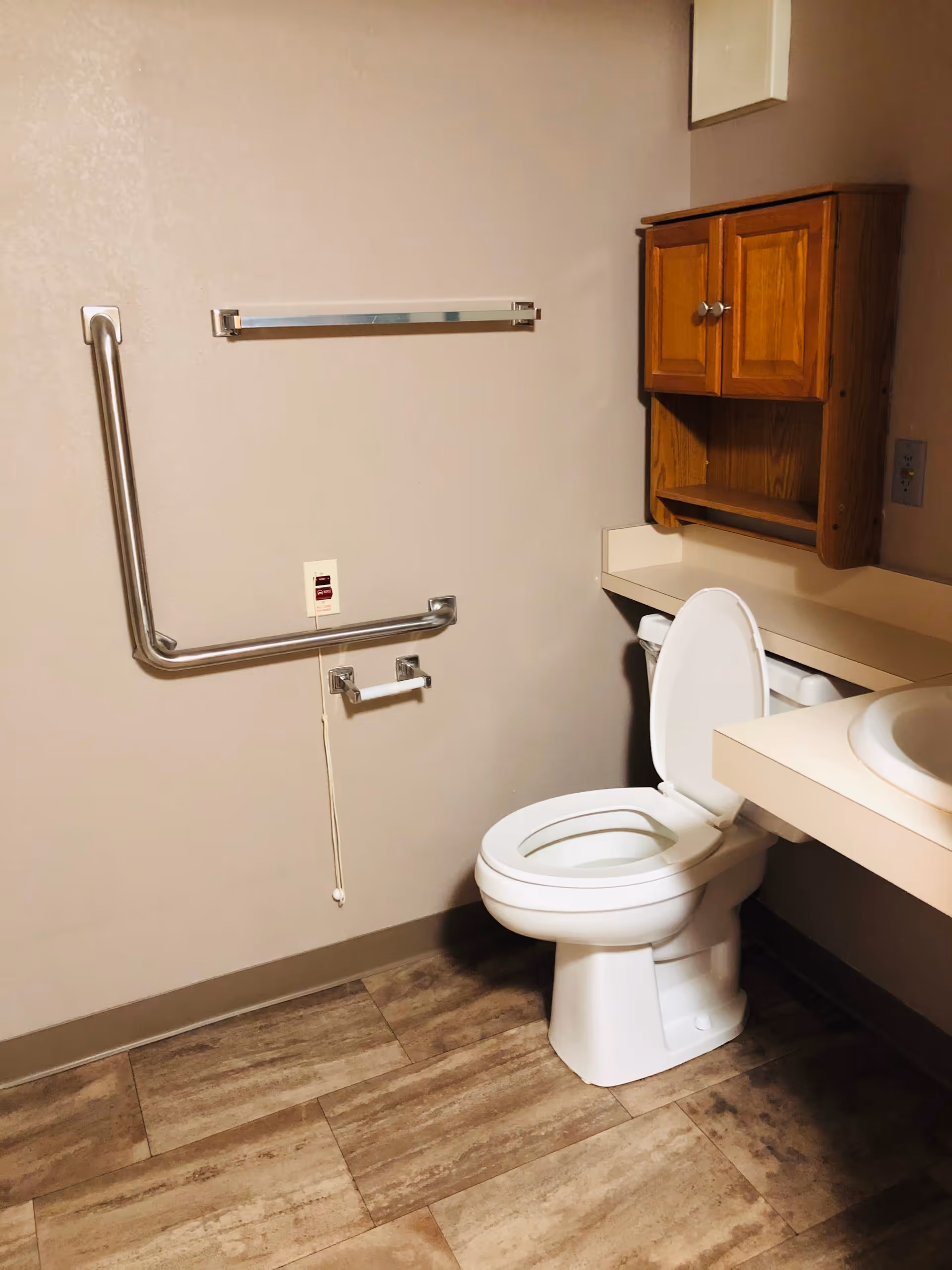 A clean bathroom with a white toilet with the lid open, a wooden cabinet mounted on the wall above the toilet, a beige countertop with a sink, a stainless steel grab bar on the wall, a toilet paper holder, and a pull cord emergency alarm. The floor has wood-like tiles.