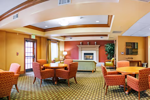 A cozy common area with multiple round wooden tables surrounded by orange patterned chairs. The room features a fireplace with a mantel decorated with small vases, a green sofa, a potted plant, and warm yellow and red walls. There is a door with glass panes and windows with curtains allowing natural light to enter.