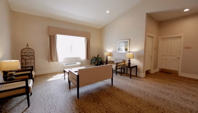 Sunlit common sitting room with a loveseat, armchairs, side tables and lamps arranged on a patterned carpet.