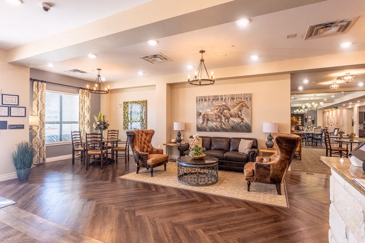 A bright and spacious living area in an assisted living facility featuring a dark leather sofa, two brown upholstered armchairs, a round coffee table with a plant centerpiece, and a painting of running horses on the wall. To the left, there is a dining area with a wooden table and chairs near a window with patterned curtains. The room has wood-patterned flooring, recessed lighting, and decorative chandeliers.