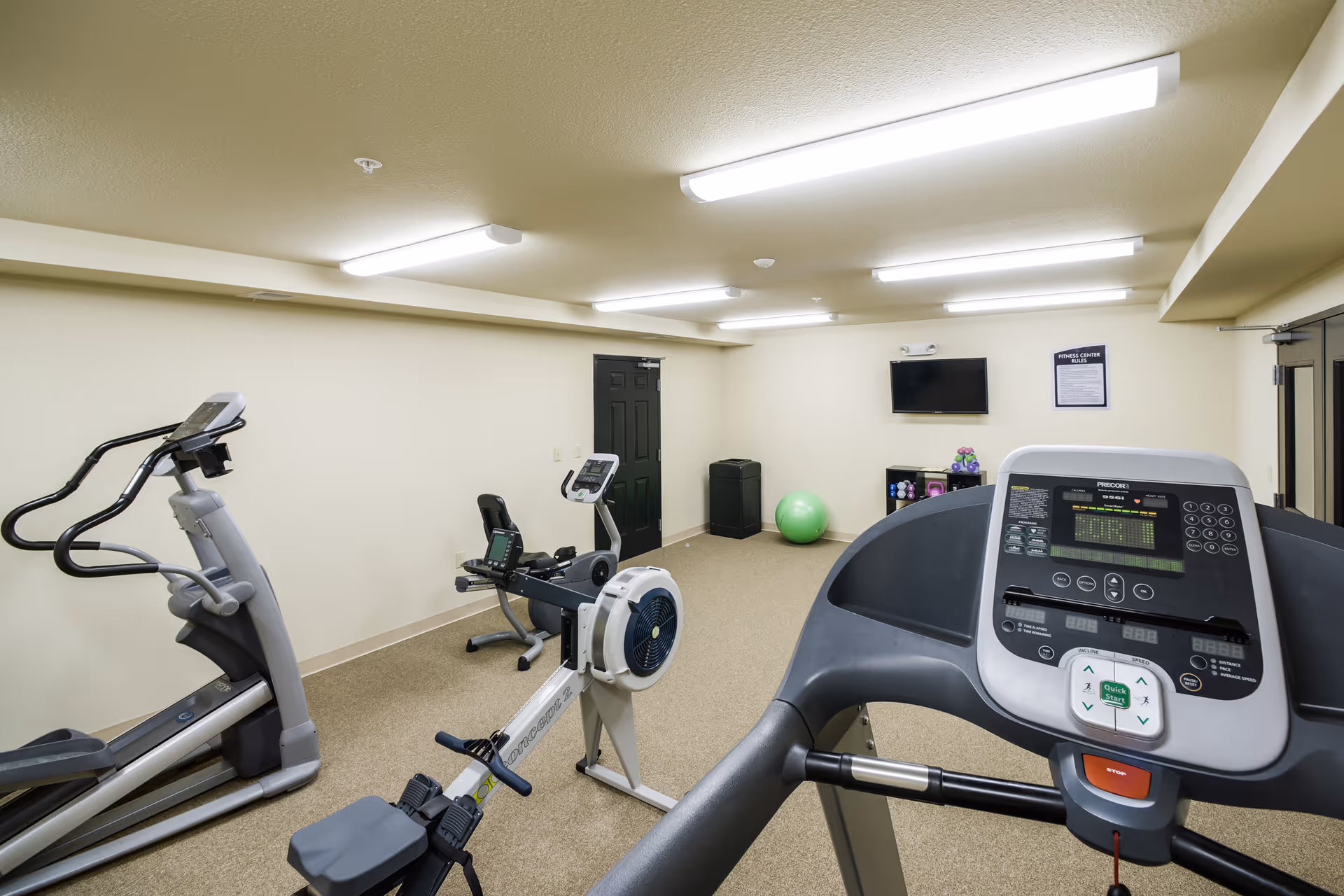 A small fitness room with exercise equipment including a treadmill, rowing machine, and elliptical trainer. The room has beige walls, fluorescent ceiling lights, a wall-mounted TV, a green exercise ball, and a small shelving unit with weights and other fitness accessories.