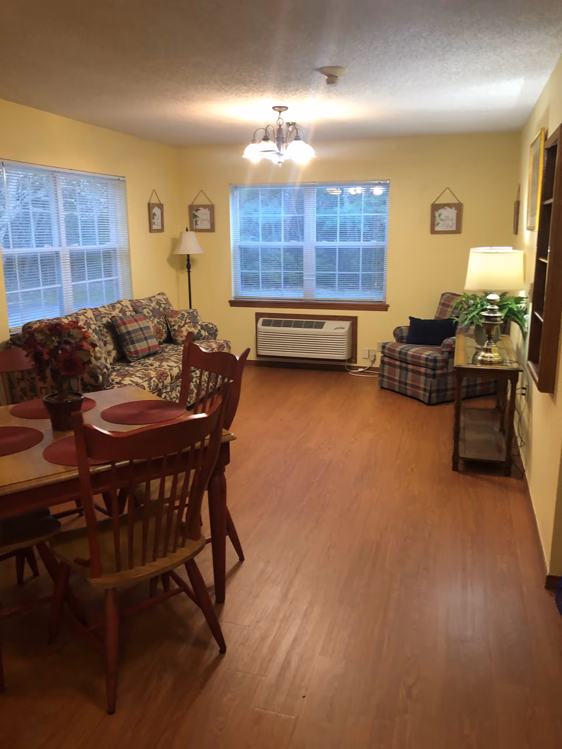A cozy living room with yellow walls and wood flooring. The room features a floral patterned sofa with plaid cushions, a wooden dining table with four chairs and red placemats, a plaid armchair, a floor lamp, a table lamp on a small wooden side table, and two large windows with blinds letting in natural light.