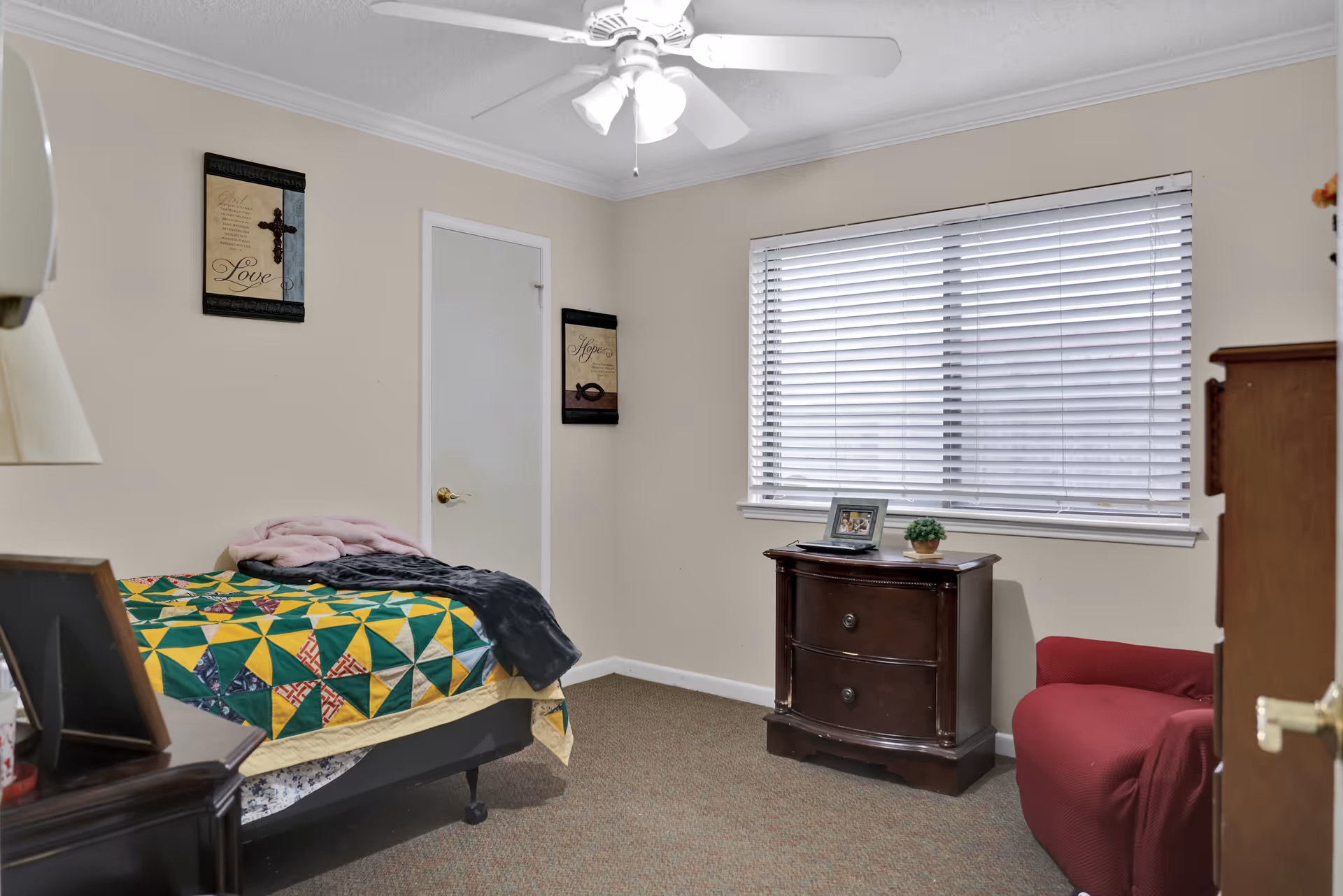 A small bedroom with a single bed covered with a colorful quilt, a dark wooden nightstand with a lamp, a dark wooden dresser with a framed photo and a small plant on top, a red armchair, a window with white blinds, and two framed wall decorations. The room has beige walls and carpeted floor.