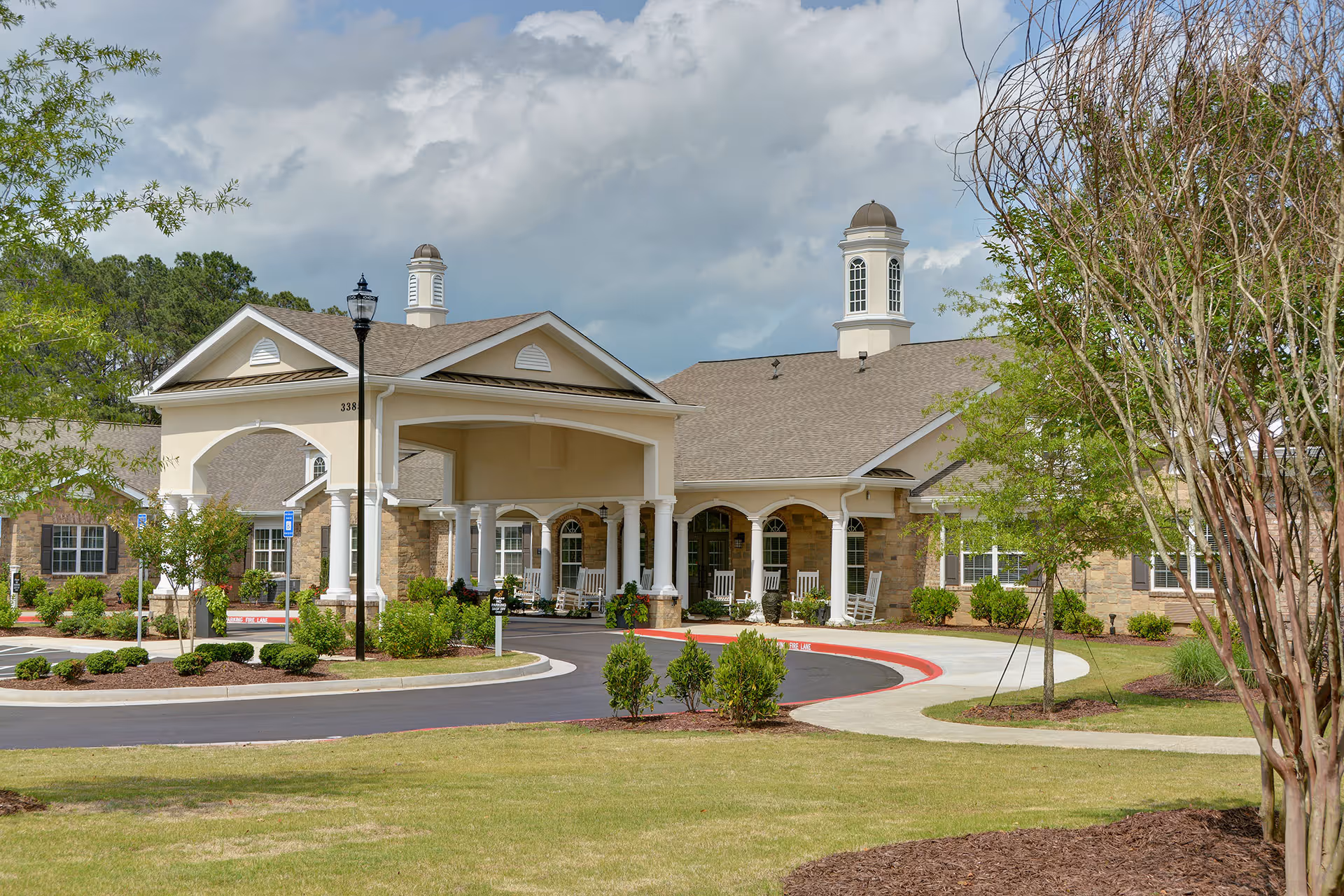 Exterior view of Benton Manor Of Stockbridge, showing a large building with a covered entrance supported by white columns, rocking chairs on the porch, manicured bushes, trees, and a curved driveway under a partly cloudy sky.