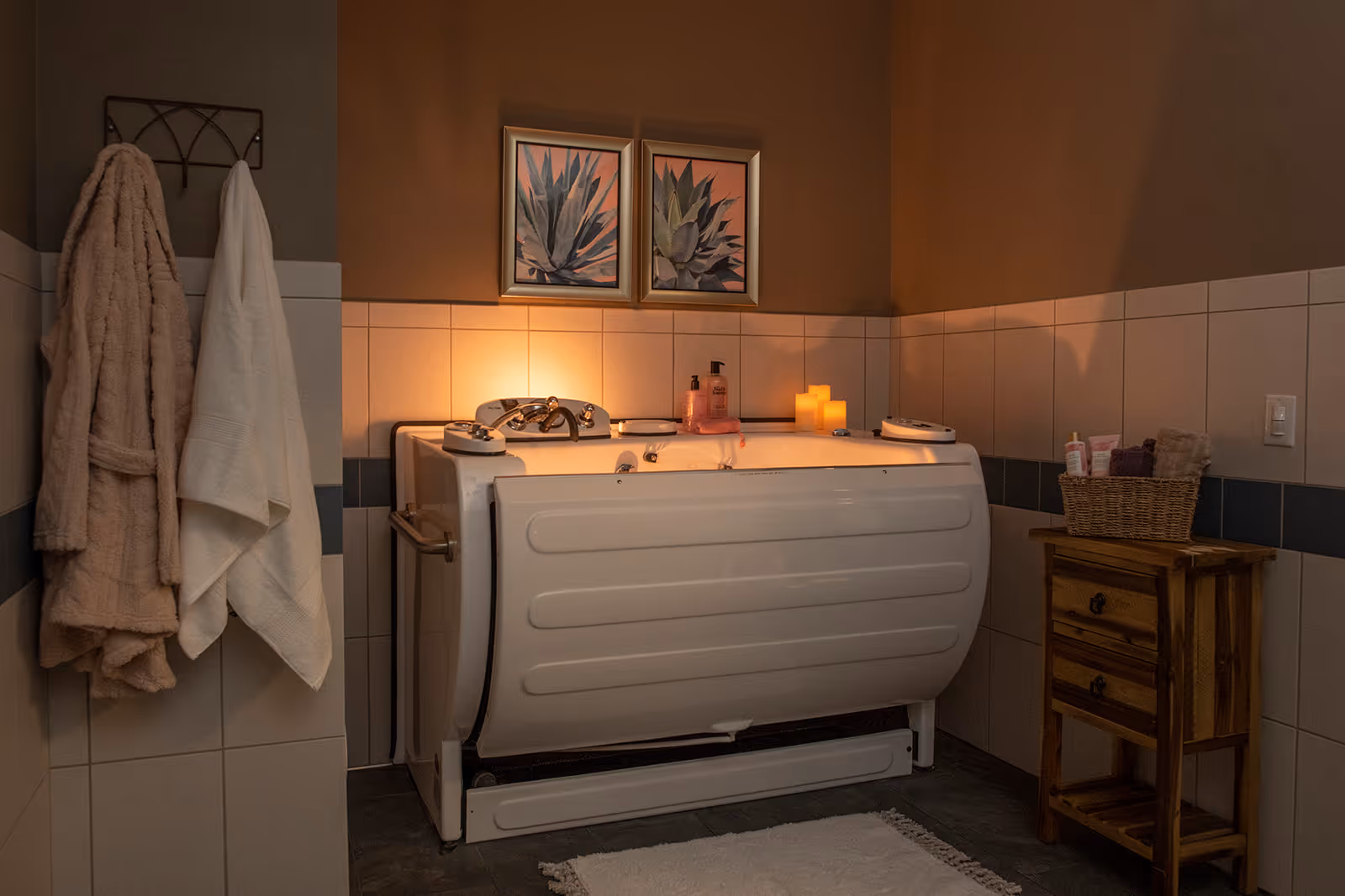 A cozy bathroom featuring a walk-in bathtub with handrails and controls on the edges. The bathtub is surrounded by tiled walls and illuminated by warm candlelight. Two framed botanical prints hang above the tub. On the left wall, two towels hang on a rack, and on the right, a wooden cabinet holds a basket with toiletries. A white bath mat is placed on the floor in front of the tub.