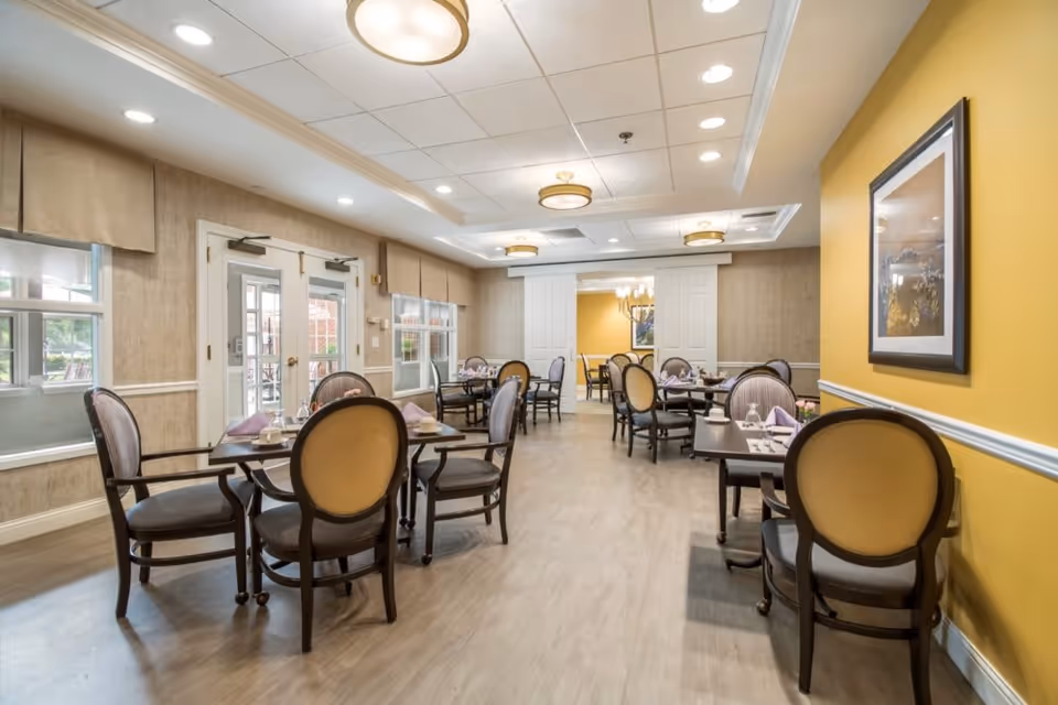 A bright and elegant dining room with multiple tables and chairs arranged neatly. Each table is set with cups, saucers, and folded napkins. The room features large windows, beige walls with a yellow accent wall, framed artwork, and ceiling lights providing warm illumination.
