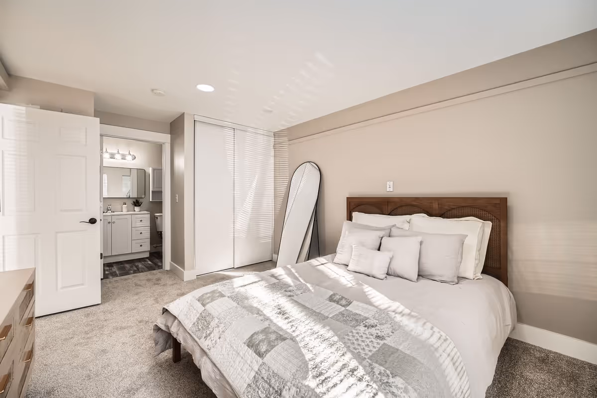 A bright bedroom with a neatly made bed featuring white and gray bedding and multiple pillows. There is a full-length mirror leaning against the wall next to a closet with sliding doors. The door to an adjoining bathroom is open, showing a vanity with a mirror and light fixtures. The room has beige walls and carpeted flooring.