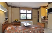 A cozy bedroom in a senior living facility featuring a bed with floral patterned bedding, a wooden dresser with a vase of yellow flowers, a window with a view outside, a wall-mounted television, and a small table in front of the bed.