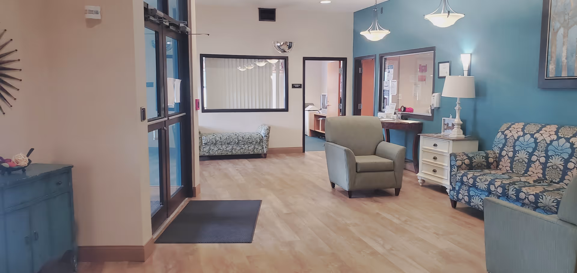 A bright and welcoming interior common area with light wood flooring, teal accent wall, and comfortable seating including armchairs and a patterned loveseat. There is a small white side table with a lamp and a bulletin board on the wall. A large window and door with glass panels are visible on the left side, and a hallway leads to other rooms in the background.