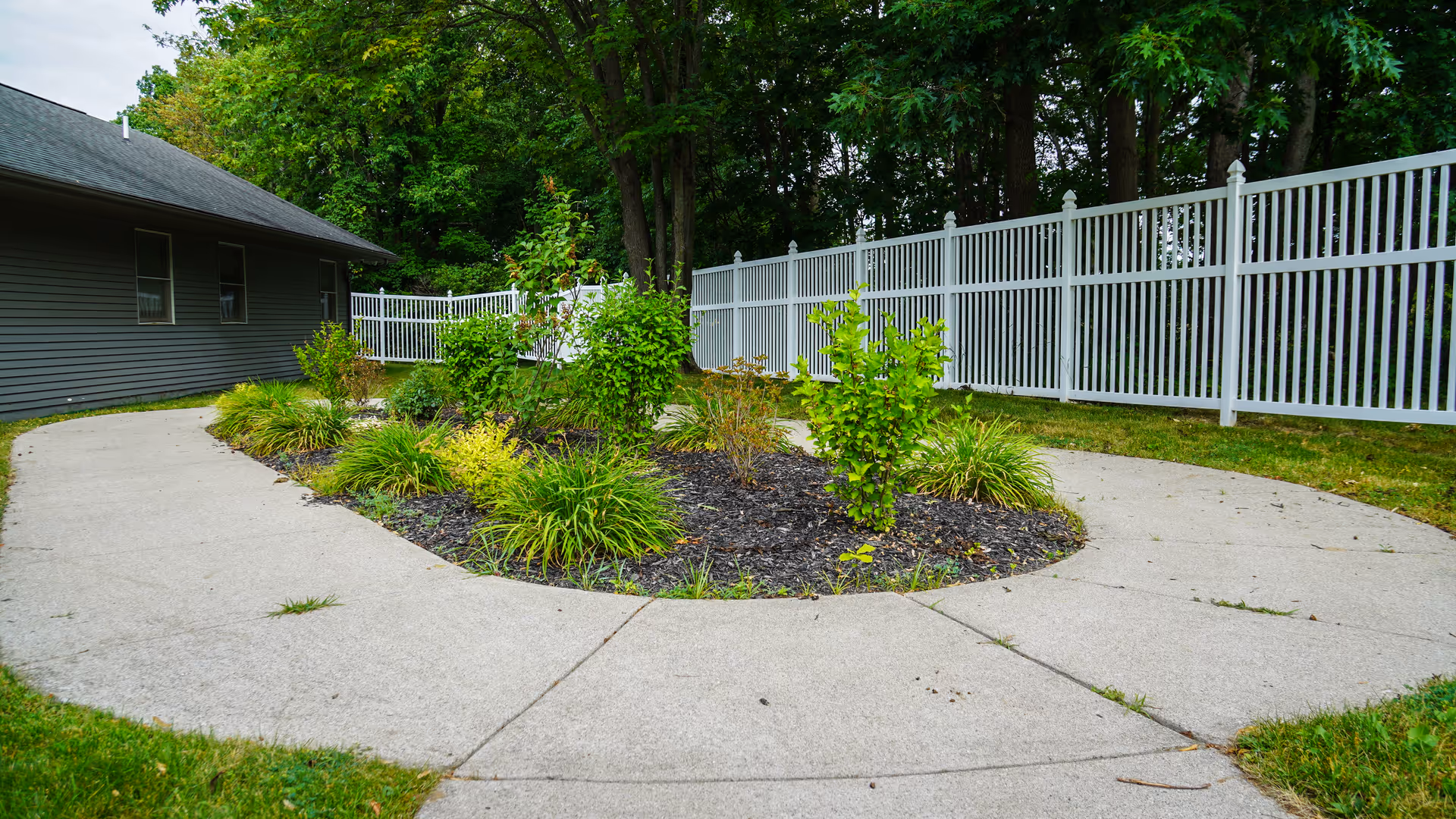 Curved concrete walkway surrounding a landscaped garden bed with various green shrubs and plants, next to a gray building with windows and a white fence in the background, bordered by trees.