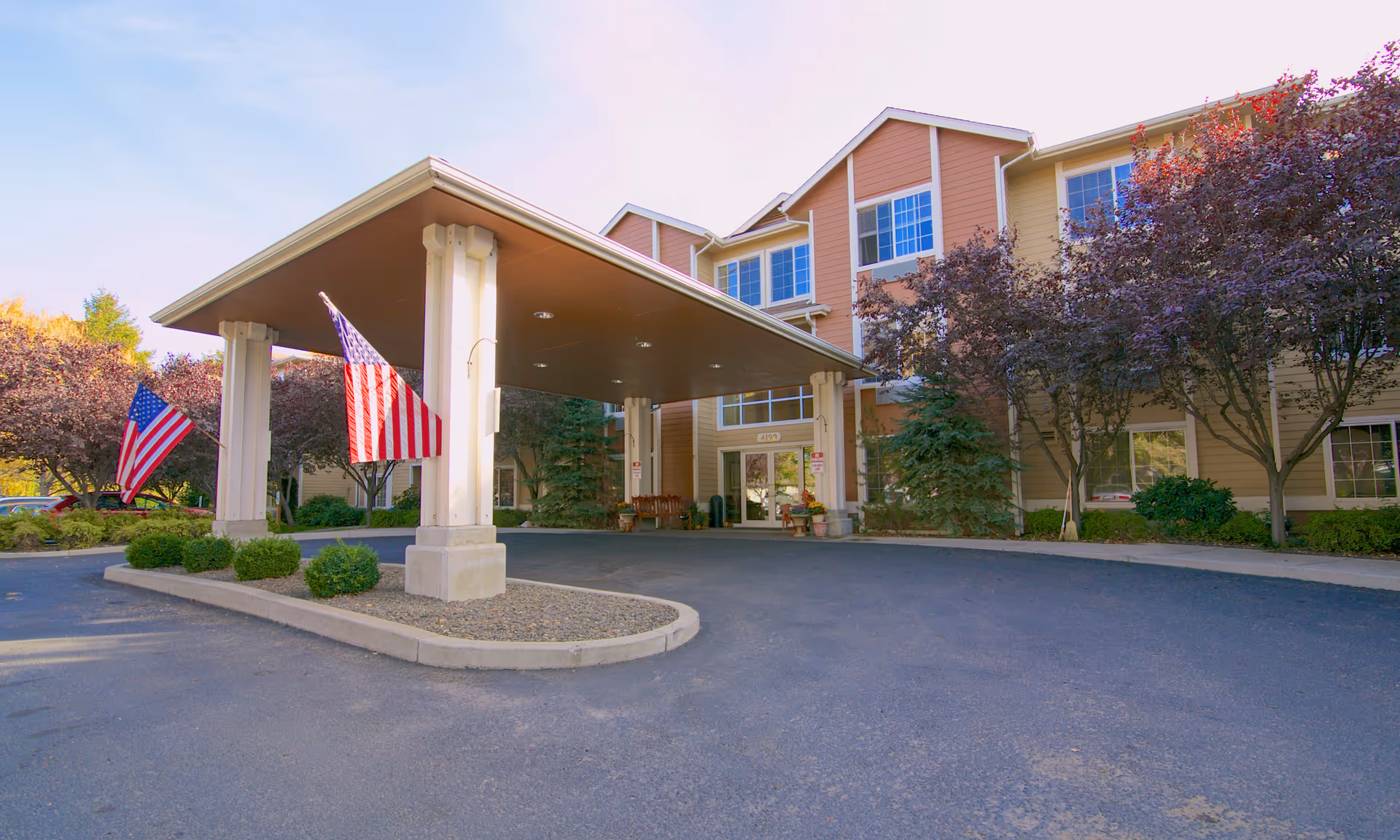Front entrance of a senior living building with a covered porte-cochere, American flags, and landscaping.