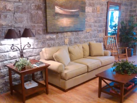 A cozy living room area with a beige sofa, a wooden coffee table with a plant on it, a wooden side table with a decorative lamp and a small plant, and a wooden rocking chair. The walls are made of stone, and there is a painting of a boat on the water hanging above the sofa. A large mirror and a potted plant are visible in the background.