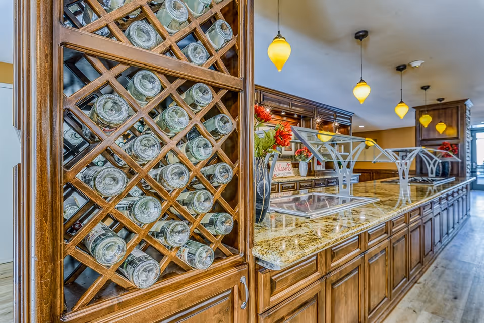 Interior view of a kitchen or dining serving area with a wooden wine rack filled with bottles on the left, a long granite countertop with metal food display stands, wooden cabinets underneath, and yellow pendant lights hanging from the ceiling.