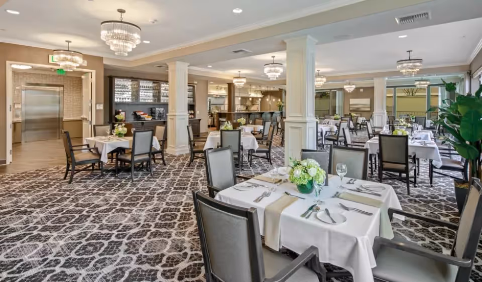 A spacious and elegant dining room with multiple tables covered in white tablecloths, each set with plates, glasses, and silverware. The room features patterned carpet, several chandeliers, large windows letting in natural light, and a serving area with a counter and bar stools in the background. There are decorative plants and artwork on the walls.