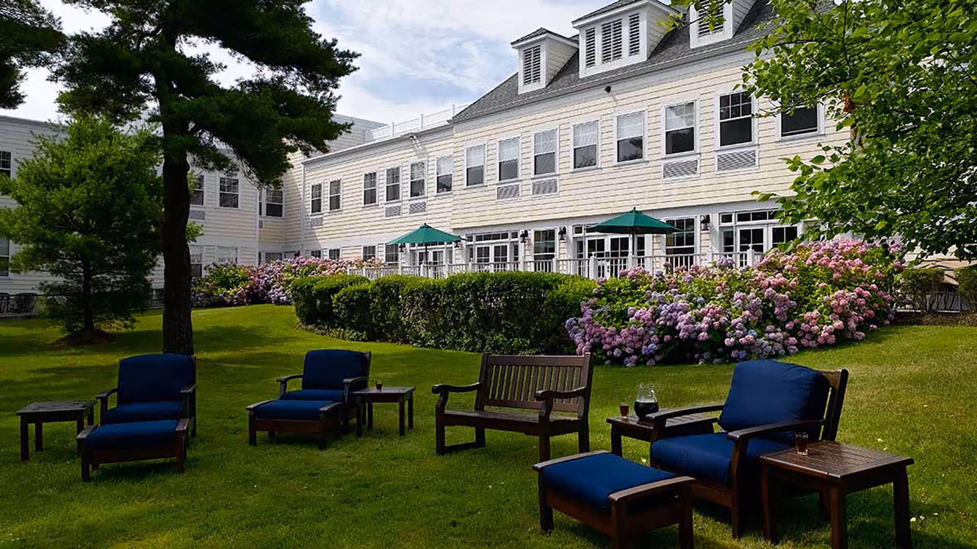 Outdoor seating area with cushioned chairs and wooden tables on a grassy lawn in front of a large white multi-story building with many windows and green umbrellas on the patio. There are trees and blooming bushes surrounding the area.