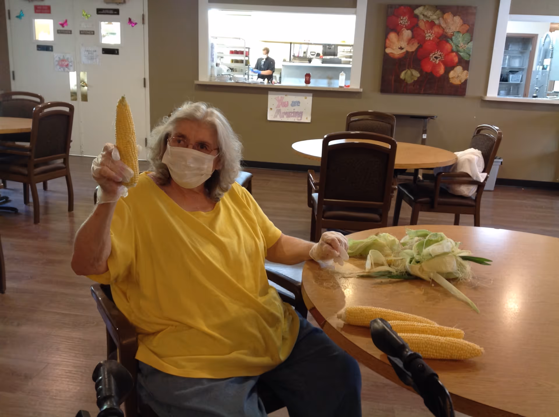 An elderly woman wearing a yellow shirt and a face mask is sitting at a round table in a dining area. She is holding an ear of corn in one hand and has peeled corn husks and two more ears of corn on the table in front of her. The room has several empty tables and chairs, a kitchen pass-through window with a person visible inside, and a floral painting on the wall.