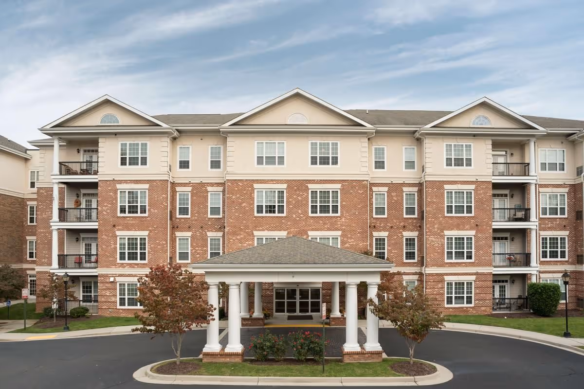 Front exterior view of a multi-story senior living facility building with a brick and beige facade, multiple windows, balconies, and a covered entrance supported by white columns. There are small trees and landscaped areas around the entrance and a circular driveway in front.