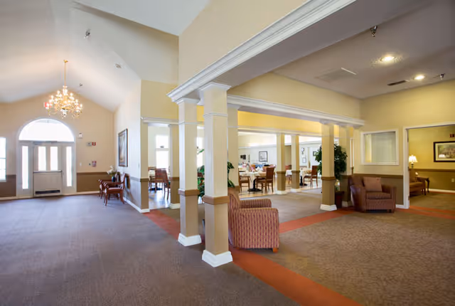 Spacious and well-lit interior common area of a senior living facility with beige walls, carpeted floors, multiple seating areas with armchairs and tables, decorative columns, and a chandelier near the entrance door with large windows.