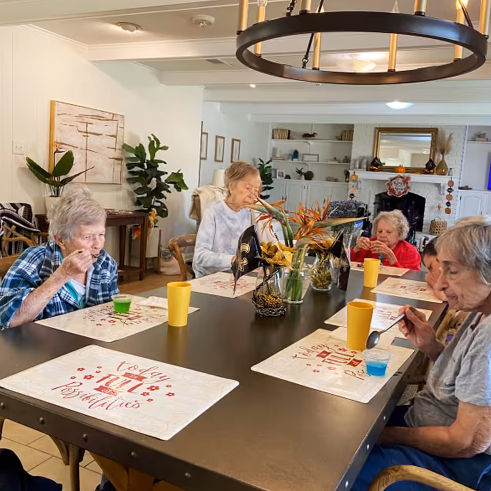 A group of elderly women sitting around a dining table in a well-lit room, eating and drinking from yellow cups and small bowls. The table is decorated with placemats and a centerpiece of flowers. The room has white walls, plants, and shelves with decorative items in the background.