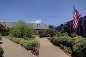 Split concrete pathways wind through a landscaped courtyard with shrubs, a wooden fence, buildings in the background, and an American flag under a clear blue sky.