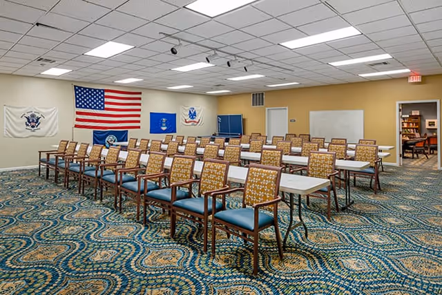 A classroom-style meeting room with rows of tables and chairs, flags on the wall, patterned carpet, and overhead fluorescent lights.