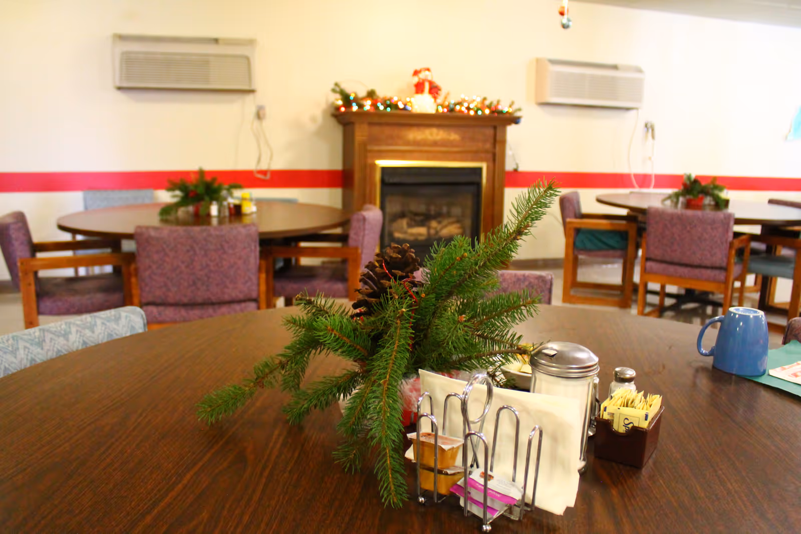 Interior view of a dining room with round wooden tables and purple cushioned chairs. A table in the foreground has a festive centerpiece with pine branches and pinecones, along with a napkin holder, sugar packets, and a blue mug. In the background, there is a wooden fireplace decorated with Christmas lights and a small Santa figure. The walls have a red stripe and two air conditioning units mounted above the fireplace.