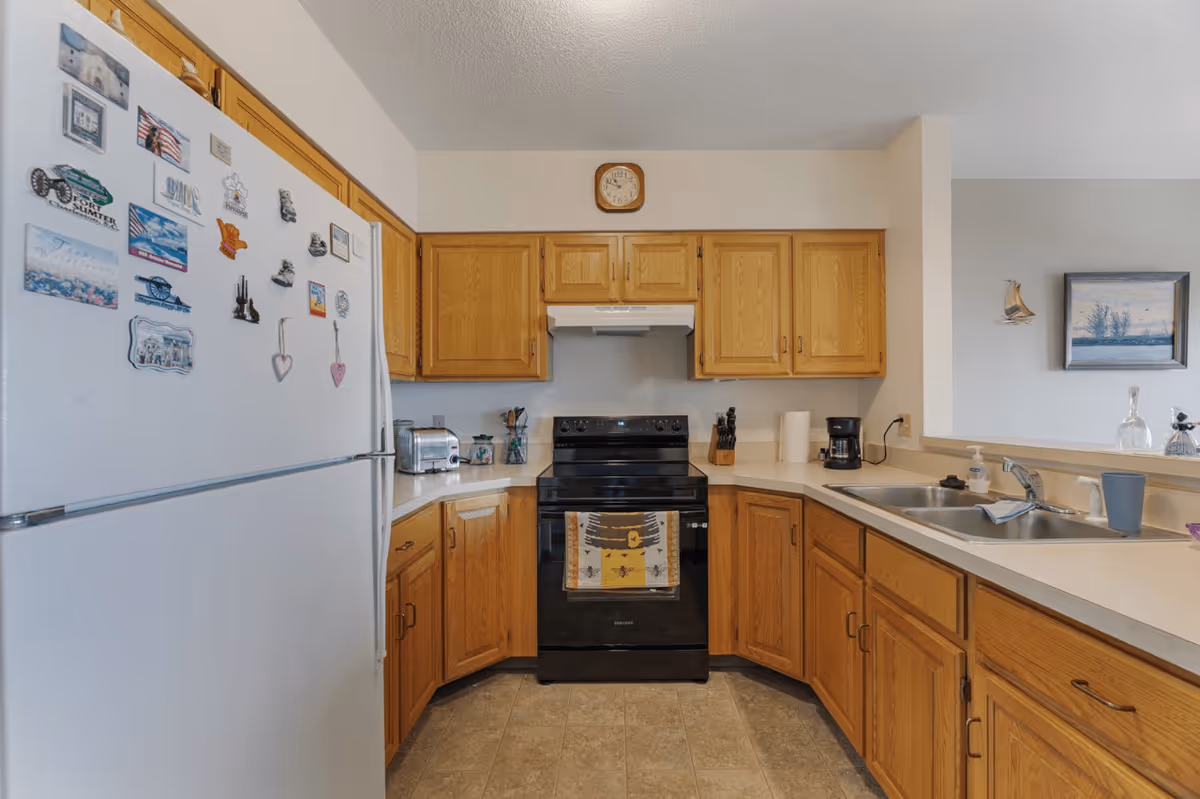 A kitchen with wooden cabinets, a white refrigerator covered with magnets, a black electric stove with a decorative towel hanging on the handle, a toaster, a coffee maker, a double sink, and a wall clock above the stove. The kitchen has beige countertops and a tiled floor.