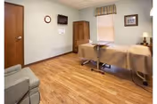 A simple, clean room with a treatment or massage table covered with a beige sheet, a wooden cabinet, a wall-mounted TV, a clock, a framed picture, a window with a striped valance, a wooden door, and a cushioned armchair on a wood floor.