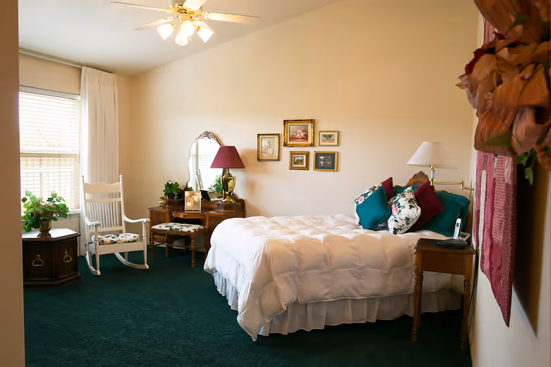 A neatly furnished bedroom with a white comforter bed, wooden nightstands and dresser, a white rocking chair, green carpet, and framed art on cream walls.