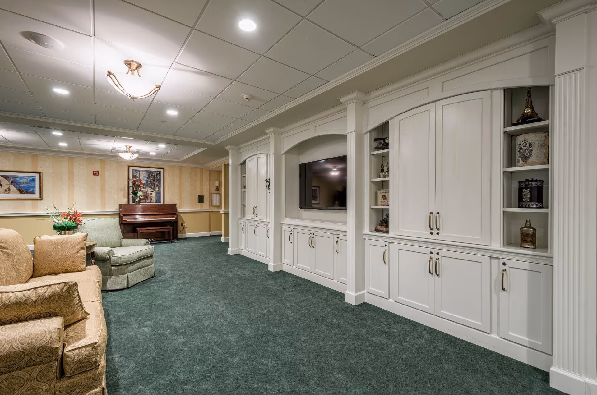 A cozy living room area in a senior living facility featuring a green carpet, beige and green upholstered armchairs and sofa, a wooden piano against the far wall, and a built-in white entertainment center with a flat-screen TV and decorative shelves.