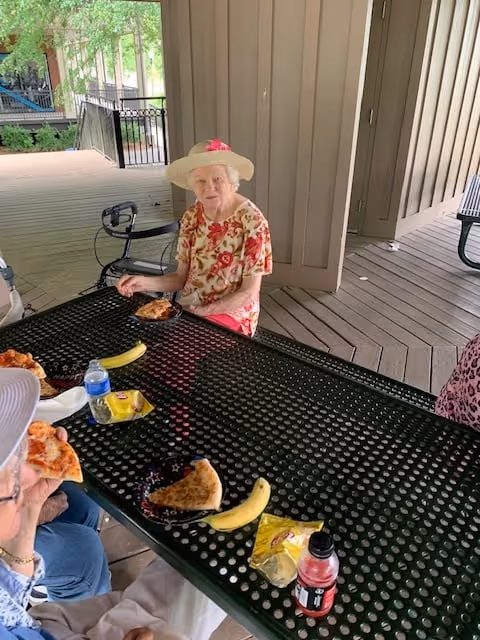 Three elderly women sitting at a black metal picnic table under a covered outdoor area. They are eating pizza and have bananas and drinks on the table. One woman is wearing a floral shirt and a wide-brimmed hat with a pink flower.