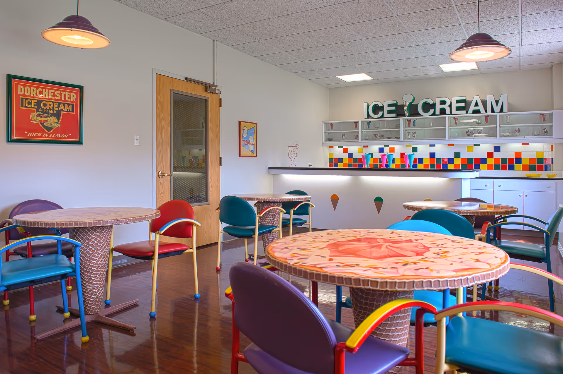 A colorful ice cream parlor-style dining area with round tables and multicolored chairs. The tables have mosaic designs resembling ice cream cones and sprinkles. The back wall features a counter with a bright multicolored tile backsplash and a large sign that reads 'ICE CREAM'. There are pendant lights hanging from the ceiling and vintage ice cream posters on the walls.
