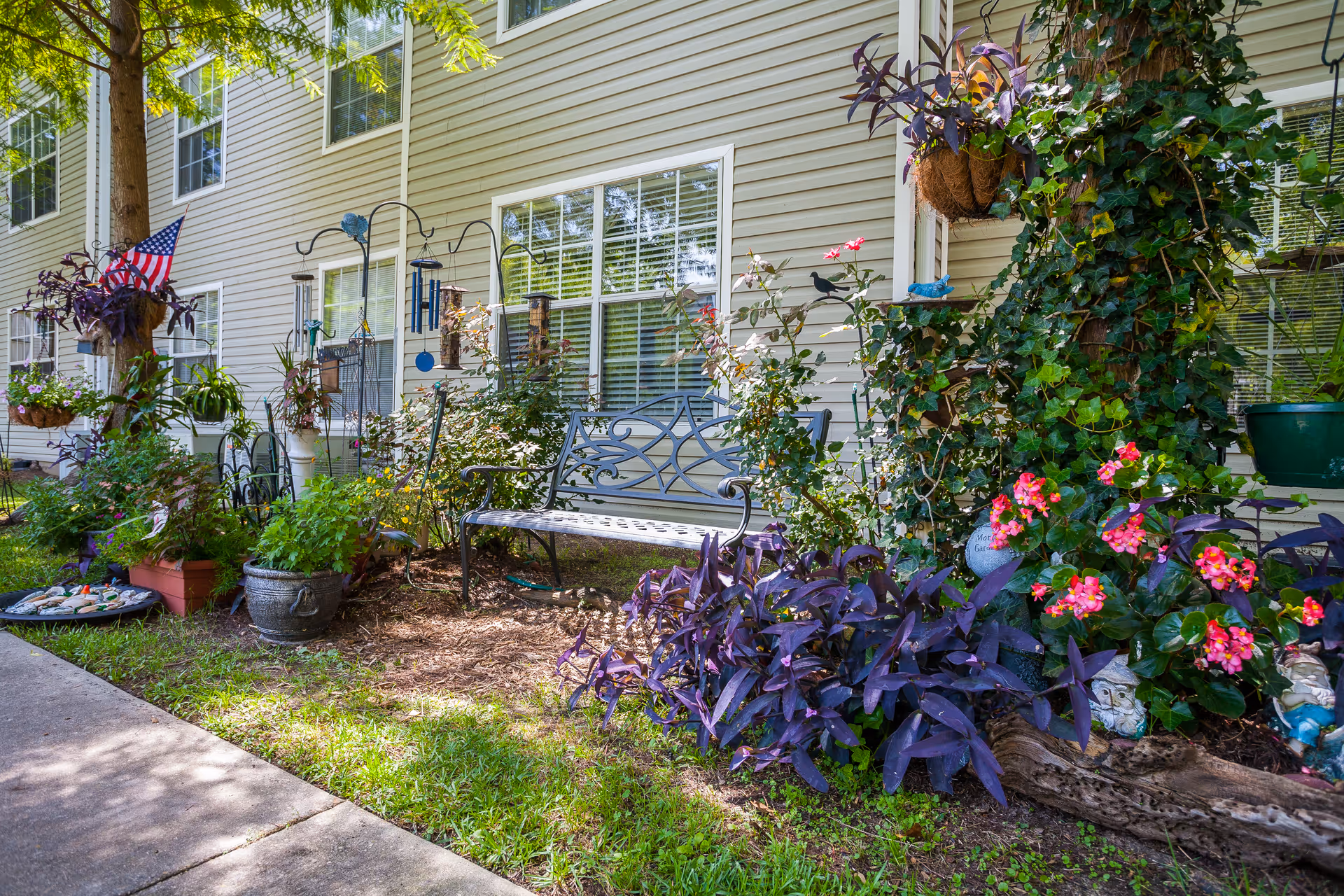 Exterior garden area alongside a senior living building with a metal bench, potted and hanging plants, and windows.