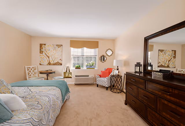 A cozy senior living bedroom with a bed covered in patterned bedding and a teal throw blanket. There is a wooden dresser with a large mirror on the right side, and a comfortable armchair with a red cushion near the window. The window has a beige valance and two small plants on the windowsill. The walls are painted light beige, and there is a decorative painting and two small mirrors on the walls. The room is carpeted and well-lit with natural light.