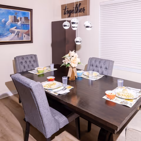 Dining room with a dark wooden table set for four, gray tufted chairs, a floral centerpiece and hanging pendant lights beneath a 'together' sign.