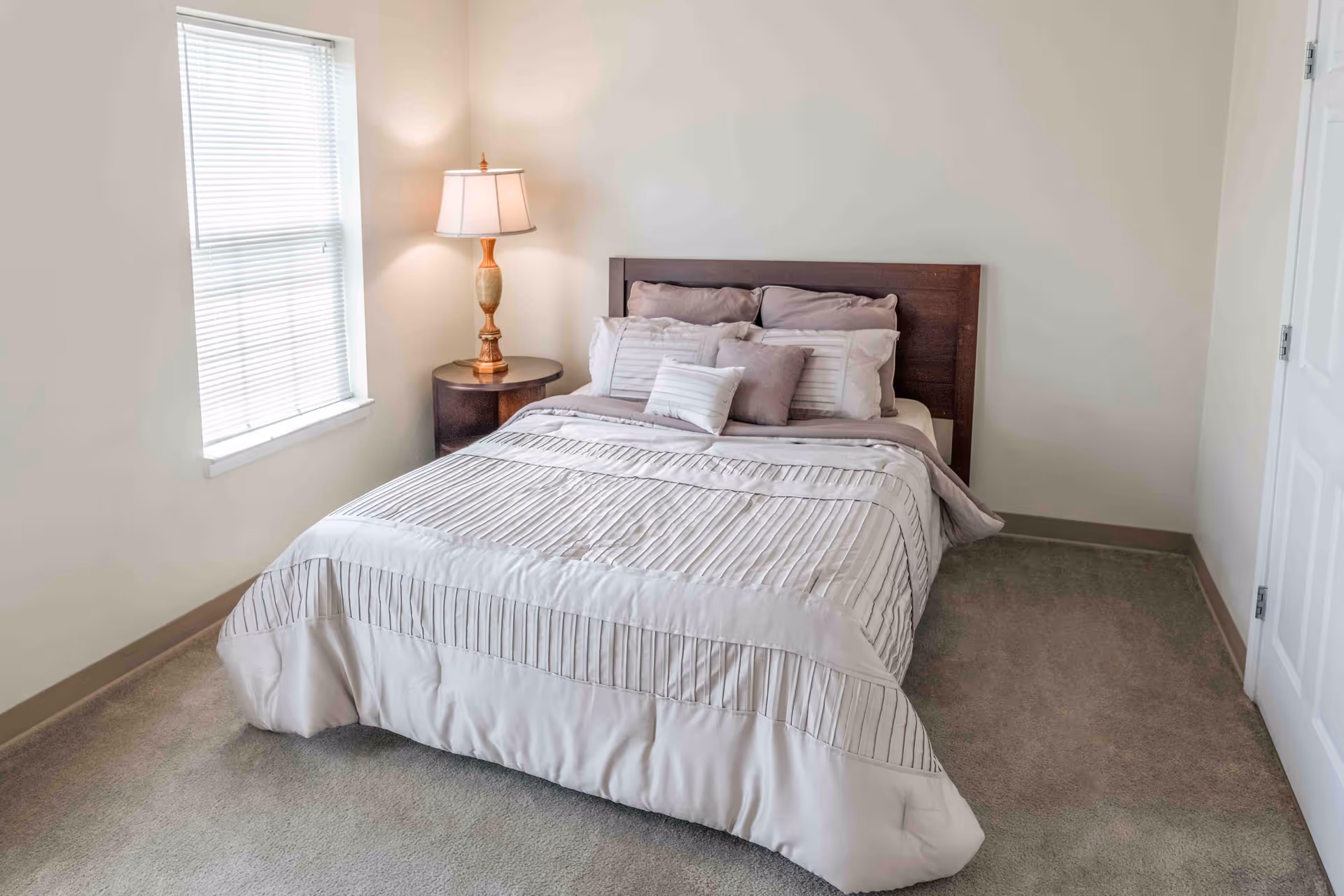 A tidy bedroom with a made bed and pillows, a wooden headboard, a round bedside table with a lamp, and a window with blinds.