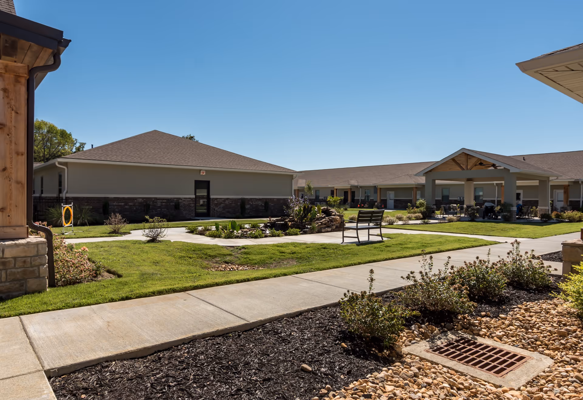 Outdoor courtyard area of Crestview Senior Living with a paved walkway, green grass, landscaped bushes, a bench, and a covered seating area attached to a single-story building under a clear blue sky.