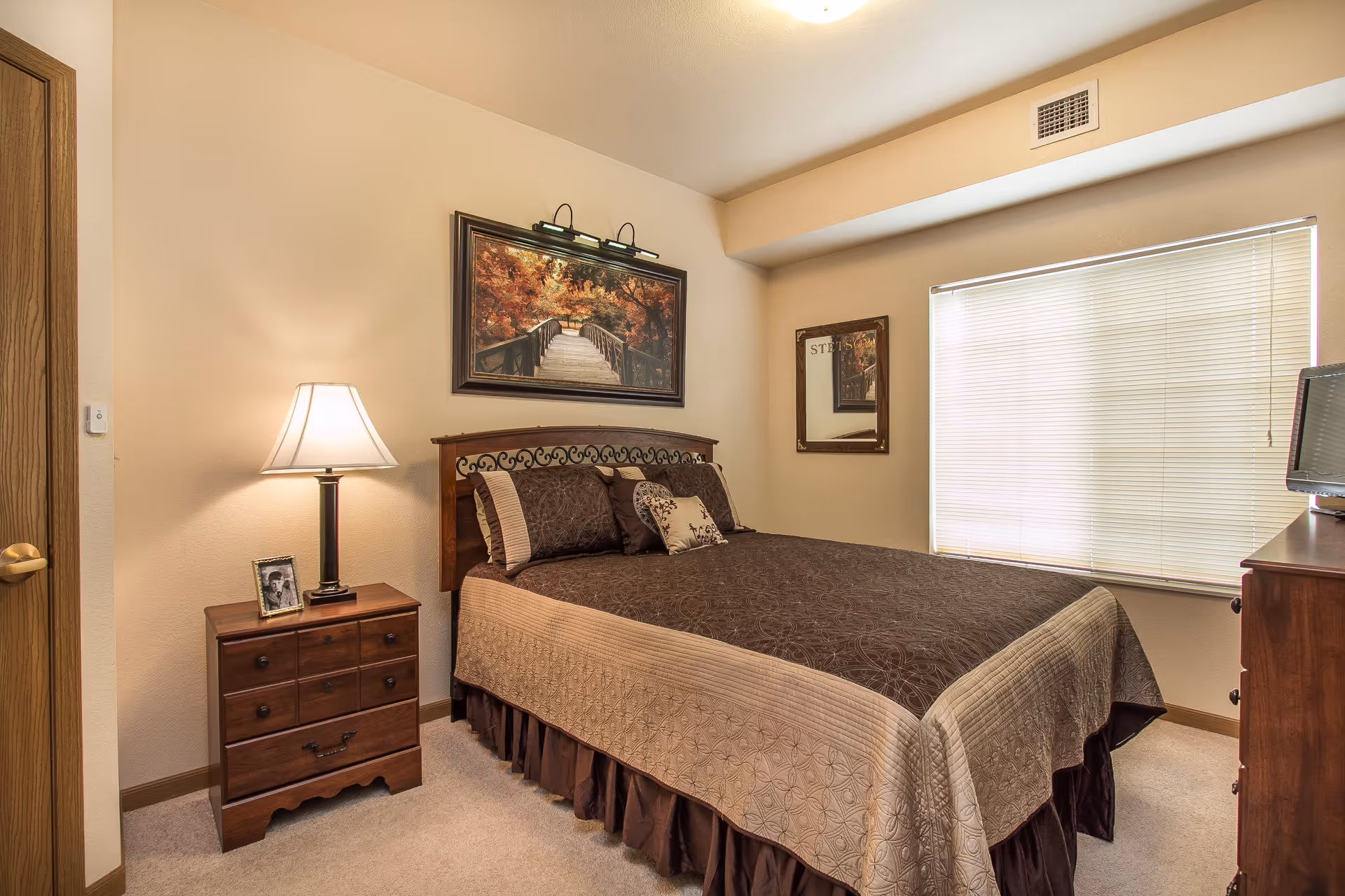 A cozy bedroom with a double bed covered in brown and beige bedding, a wooden nightstand with a lamp and a framed photo, a dresser with a TV on top, a window with blinds, and wall art including a painting of a bridge in autumn and a mirror.