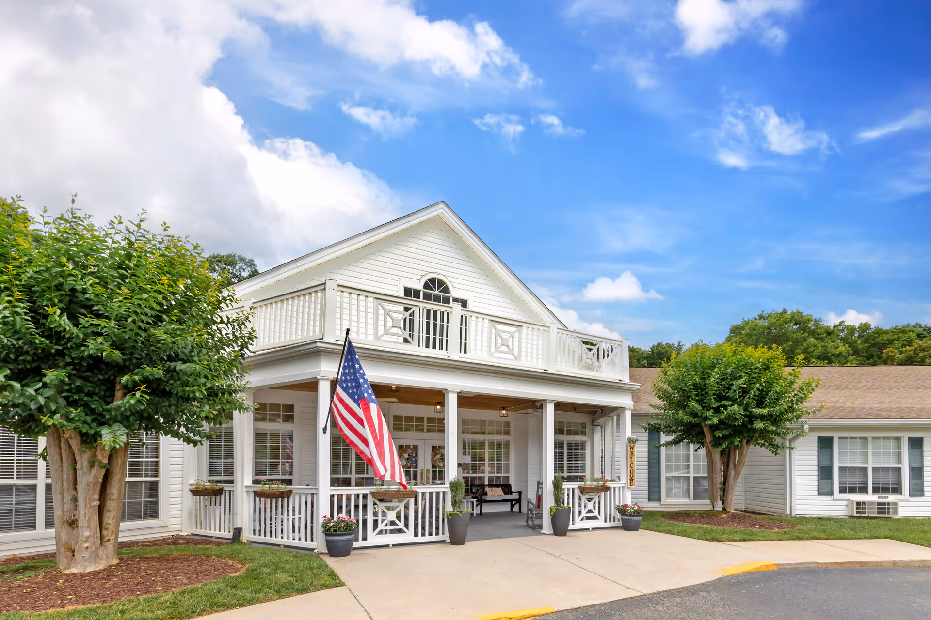 Front exterior view of a white two-story building with a porch, an American flag, potted plants, and two trimmed trees on either side under a partly cloudy blue sky.